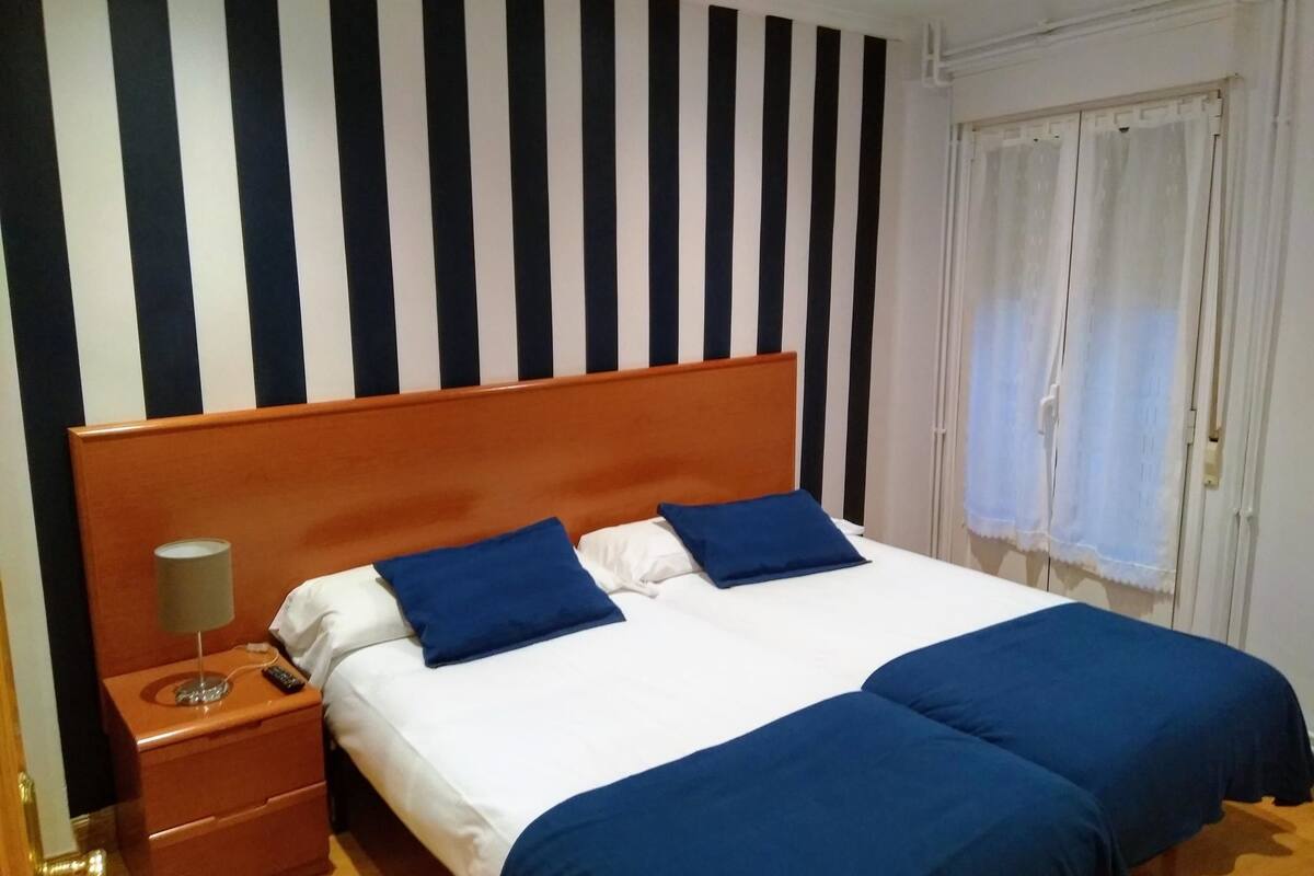 A hotel room with a large bed, two pillows, a nightstand with a lamp, and a window with curtains.