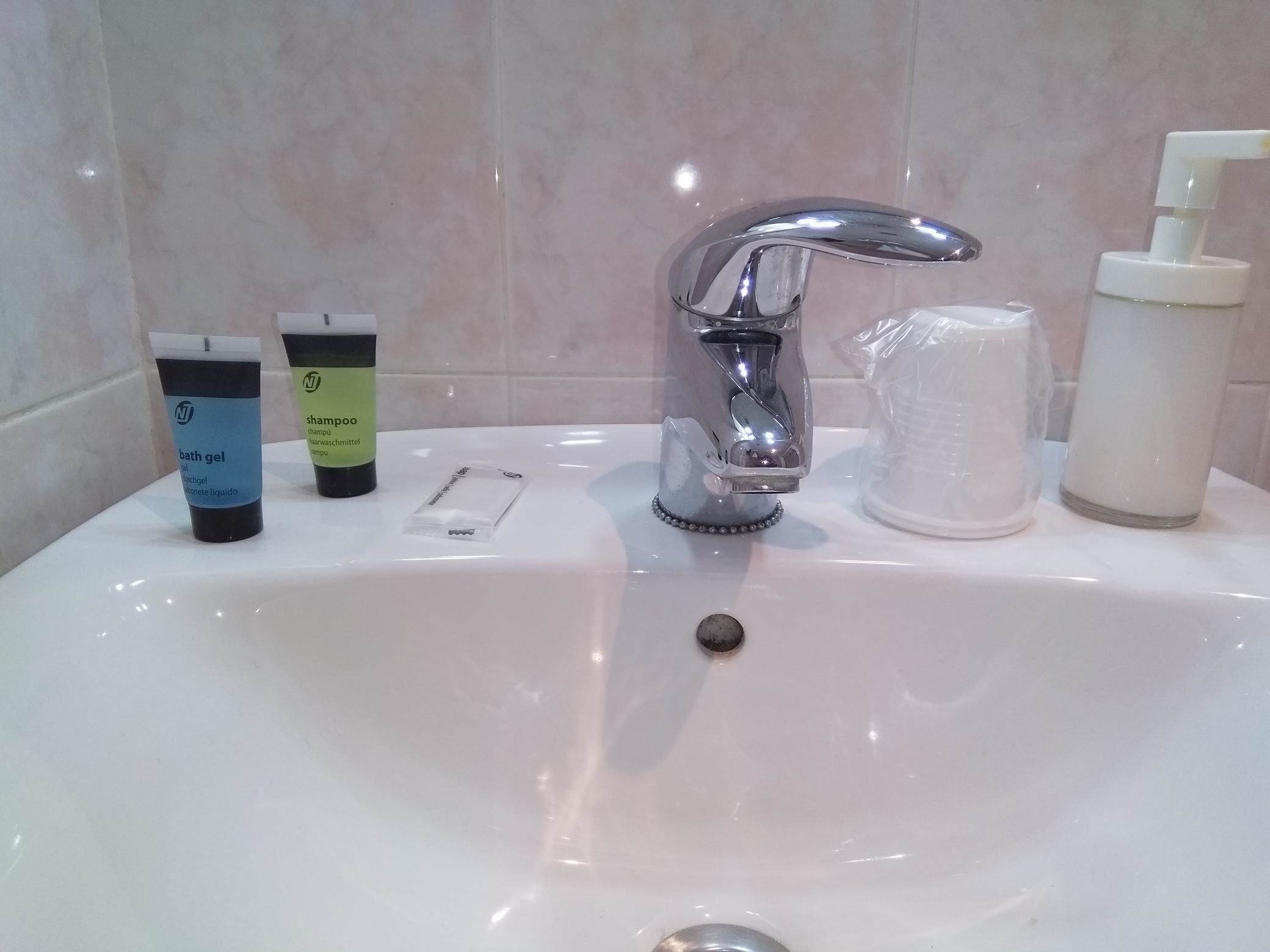 A bathroom sink with a chrome faucet, a bottle of shampoo, a tube of bath gel, and a dispenser of liquid soap.