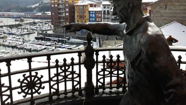 A statue of a man pointing to a marina with colorful buildings in the background. - Aldatzeta Ostatua (Bermeo)