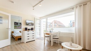 Une pièce lumineuse comprenant un lit, un bureau et donnant sur les bâtiments environnants.
