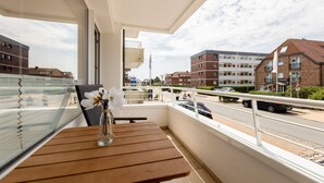Un balcon avec une table en bois et un vase de fleurs, donnant sur une rue bordée d’immeubles et de voitures.