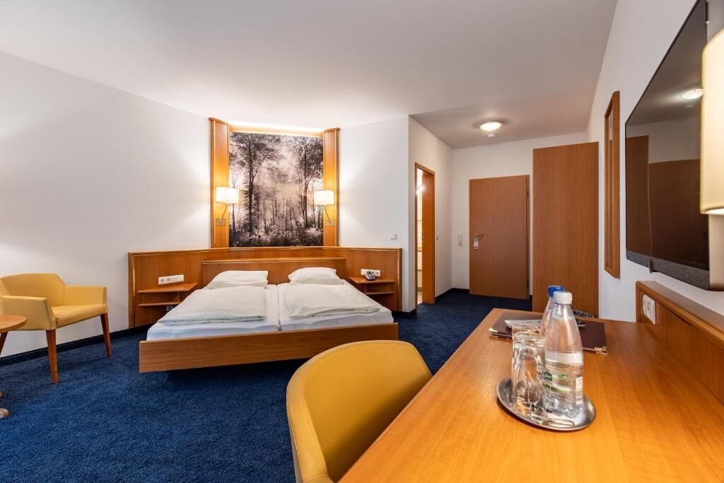 A hotel room with a bed, a desk with a water bottle, a chair, and a painting on the wall.