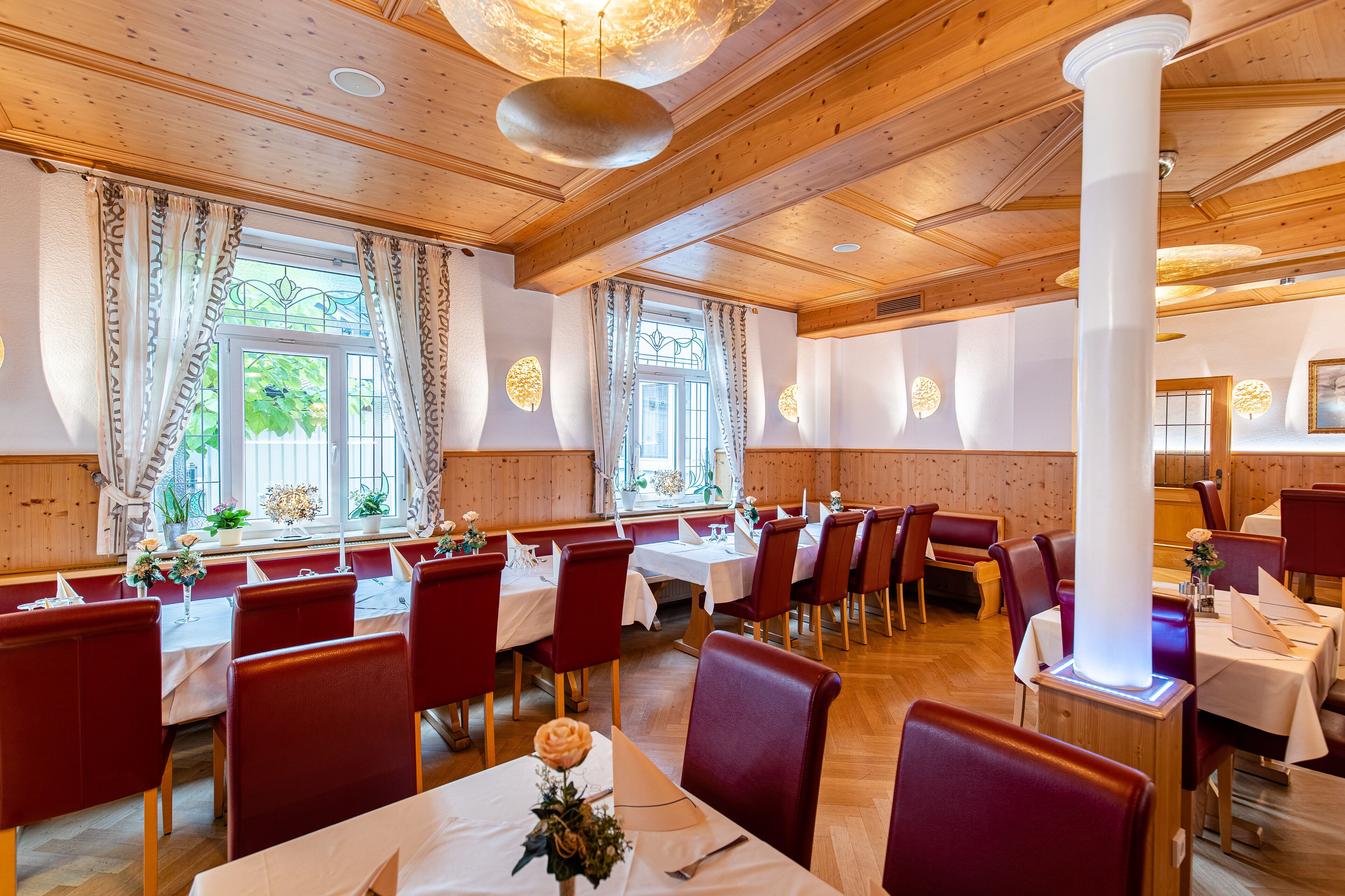 A restaurant with wooden ceiling beams, red upholstered chairs, and tables set with white tablecloths and floral centerpieces.