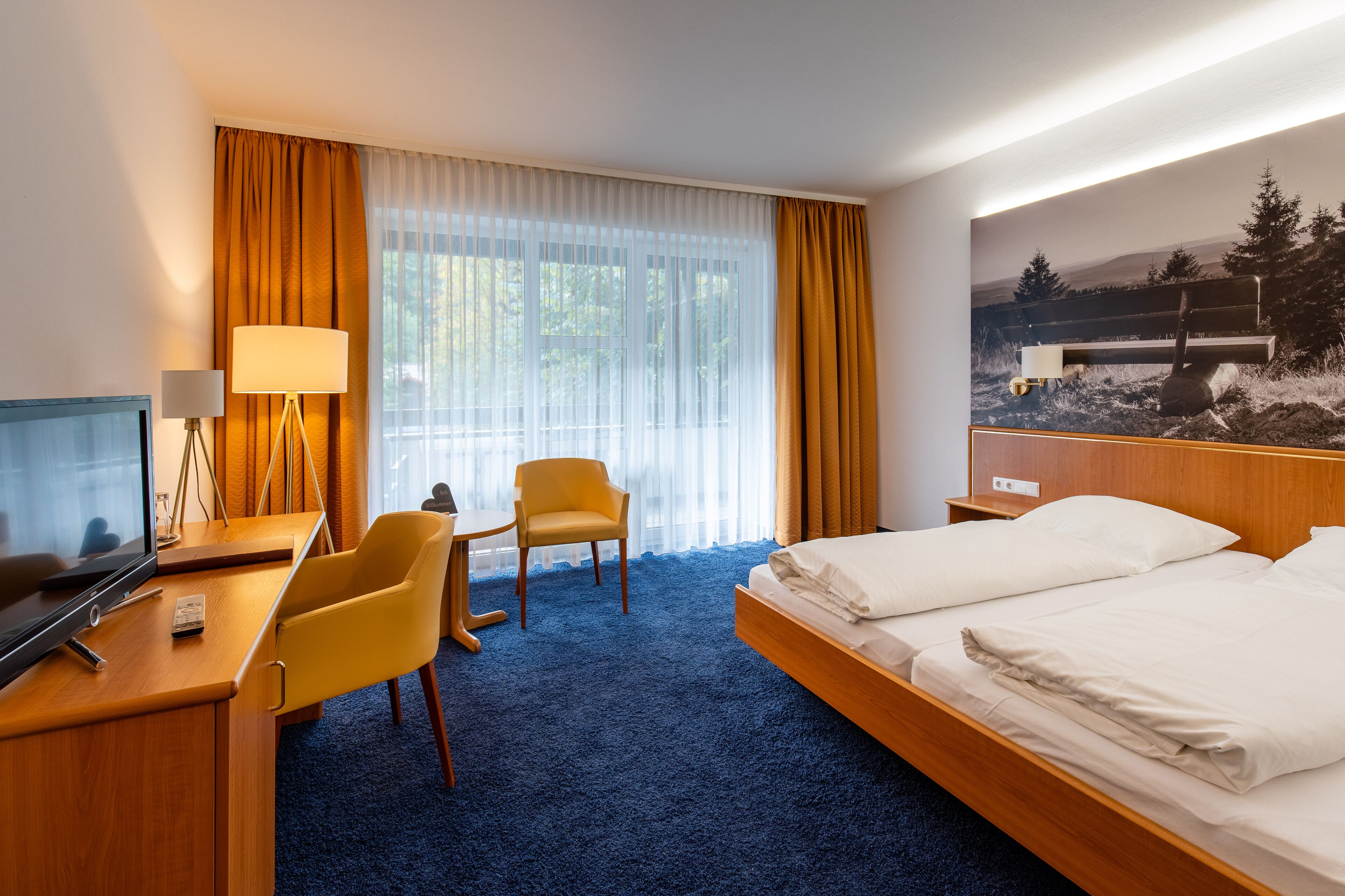 A hotel room with a bed, a desk with a chair, a TV, and a large window with curtains.