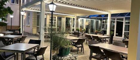 Un área de patio al aire libre con mesas y sillas, una estructura de pérgola y una jardinera de piedra.