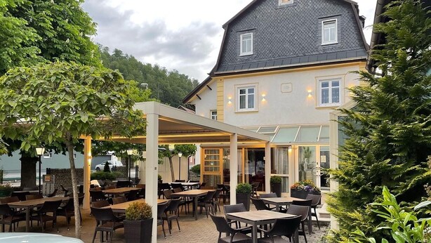 An outdoor patio area with tables and chairs, surrounded by greenery and a building with large windows.