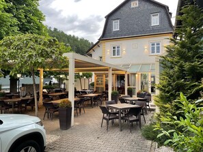 An outdoor patio area with tables and chairs, surrounded by greenery and a building with large windows. - Hotel - Gasthof Hüttensteinach (Sonneberg)