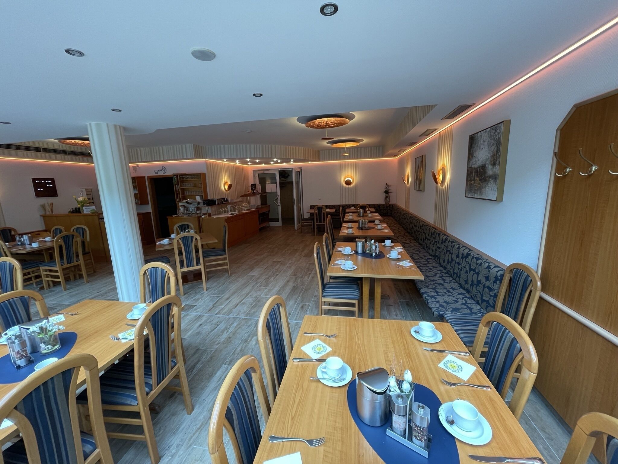 Un grand espace dédié à la salle à manger, avec de longues tables et des bancs, une cuisine à l’arrière-plan et un design moderne au niveau du plafond.