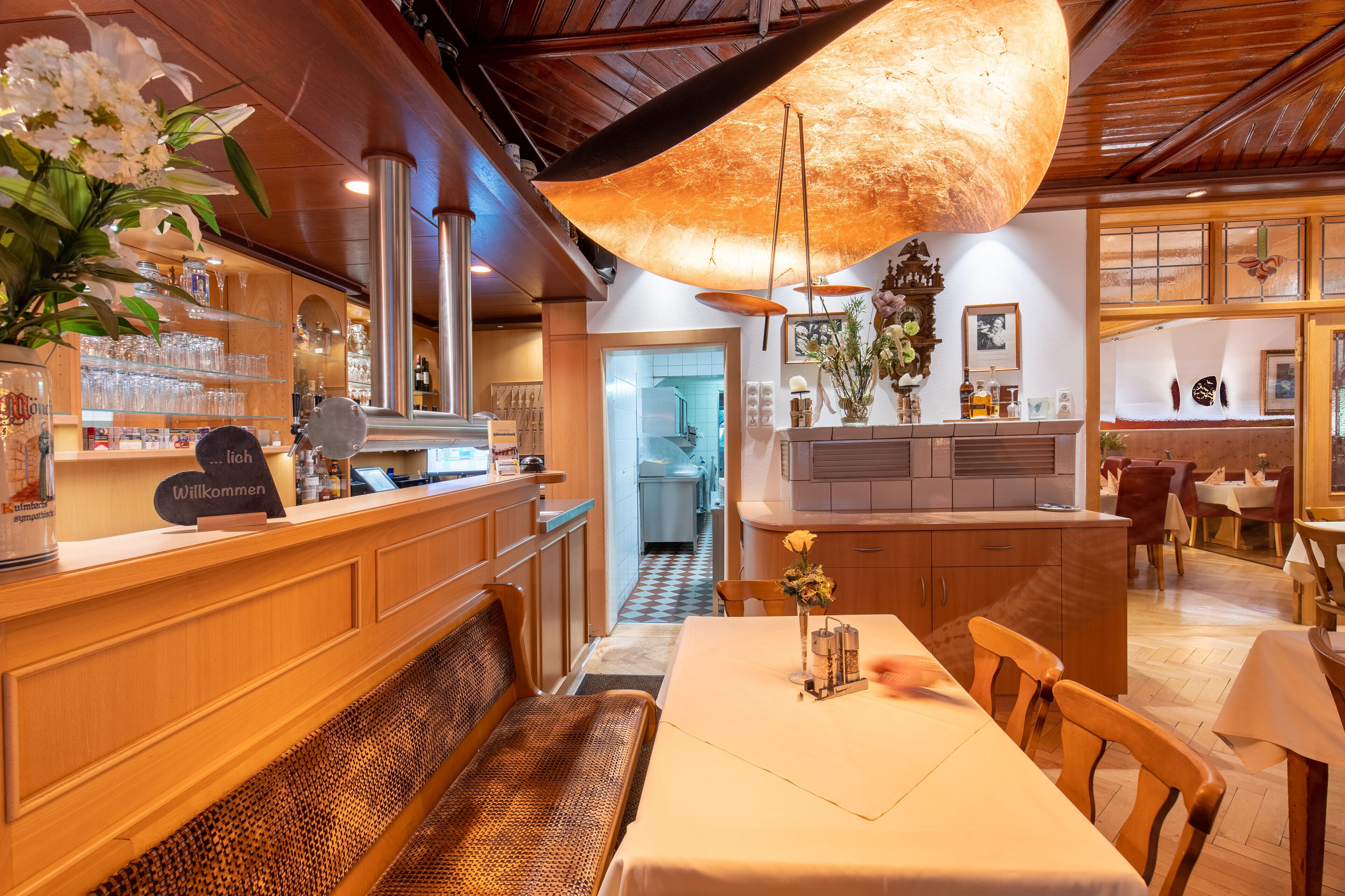 Un restaurante con muebles de madera, una lámpara colgante grande y una zona de recepción con un cartel que dice 'Willkommen'.