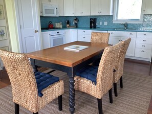 Un comedor con una mesa de madera y sillas tejidas, una cocina al fondo con armarios blancos y un salpicadero azulejado, y un ventanal que proporciona luz natural.