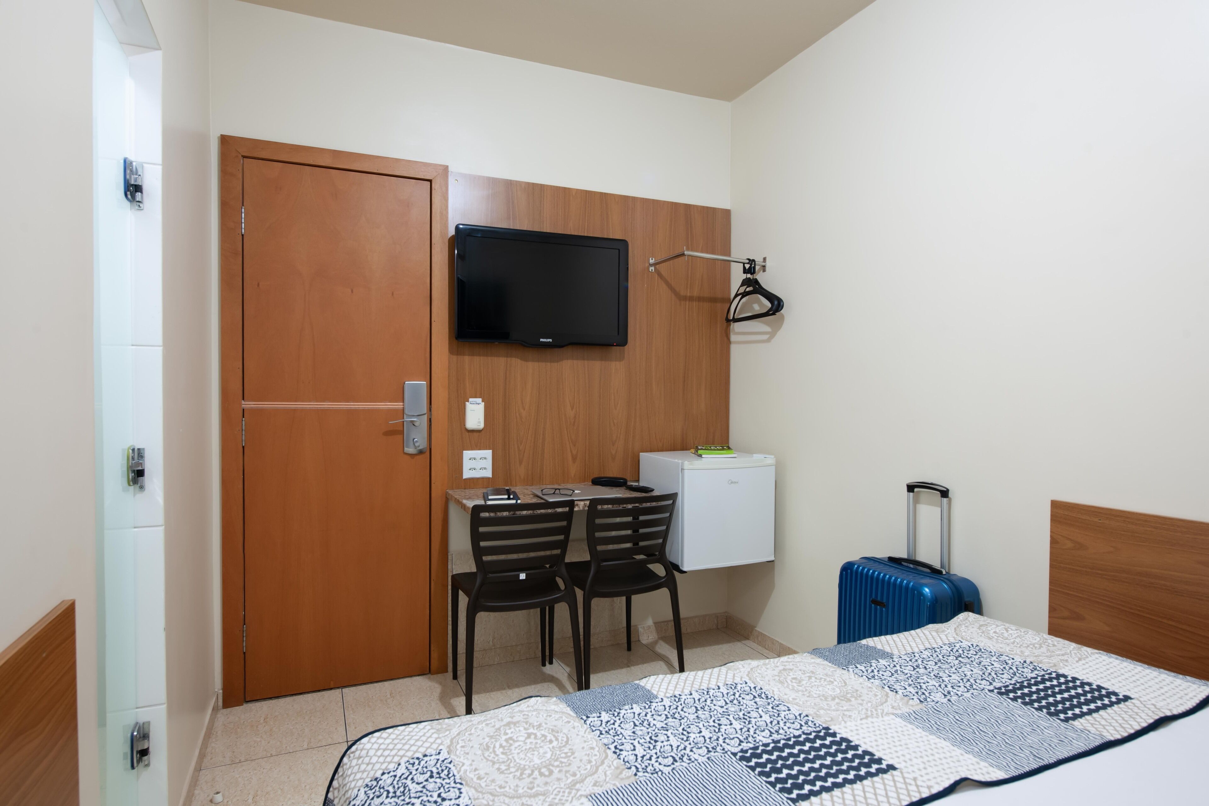 A hotel room with a bed, a desk with two chairs, a television mounted on a wooden panel, a small refrigerator, and a suitcase.