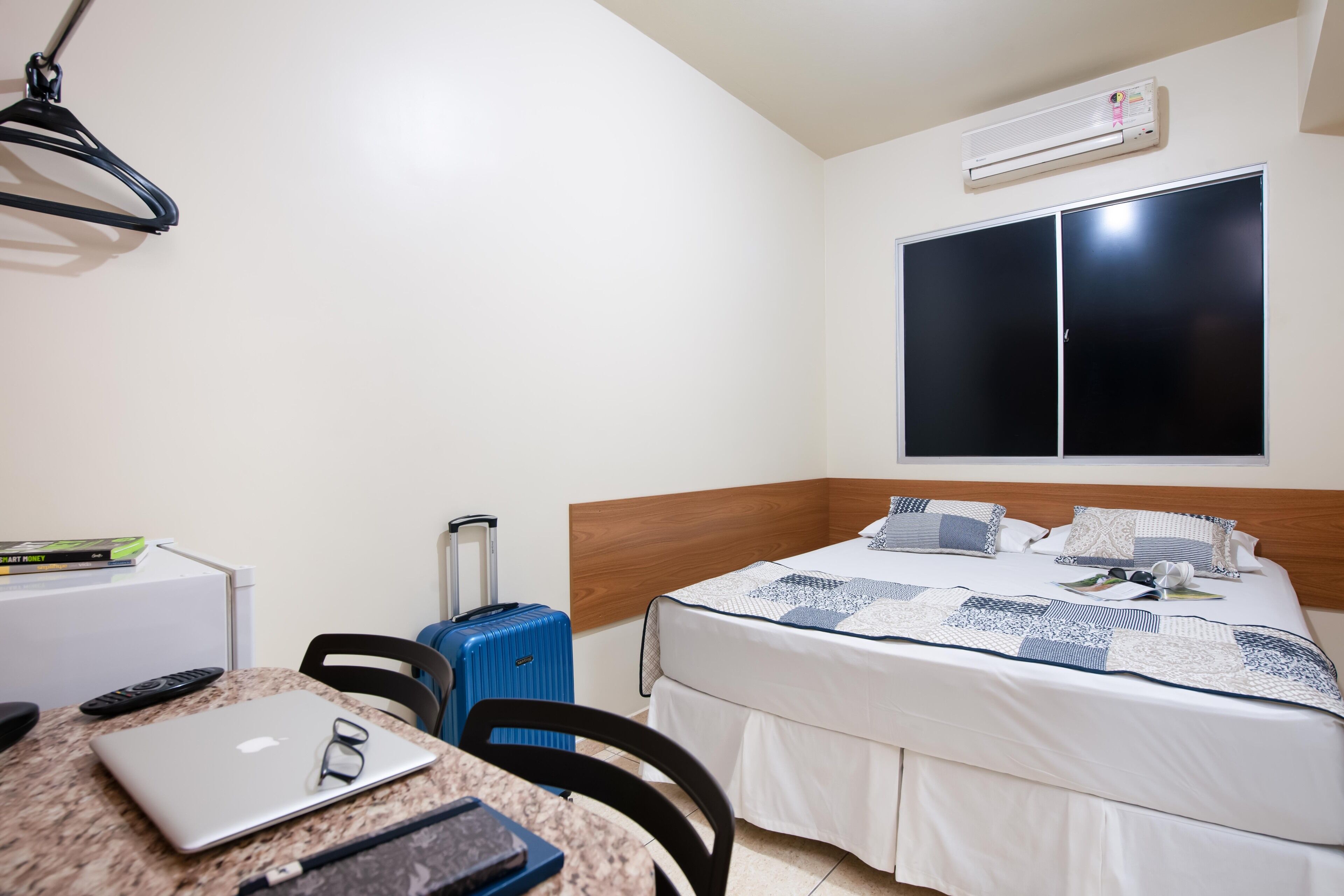 Una habitación de hotel con una cama, un escritorio con una computadora portátil, una maleta, una silla y una ventana.