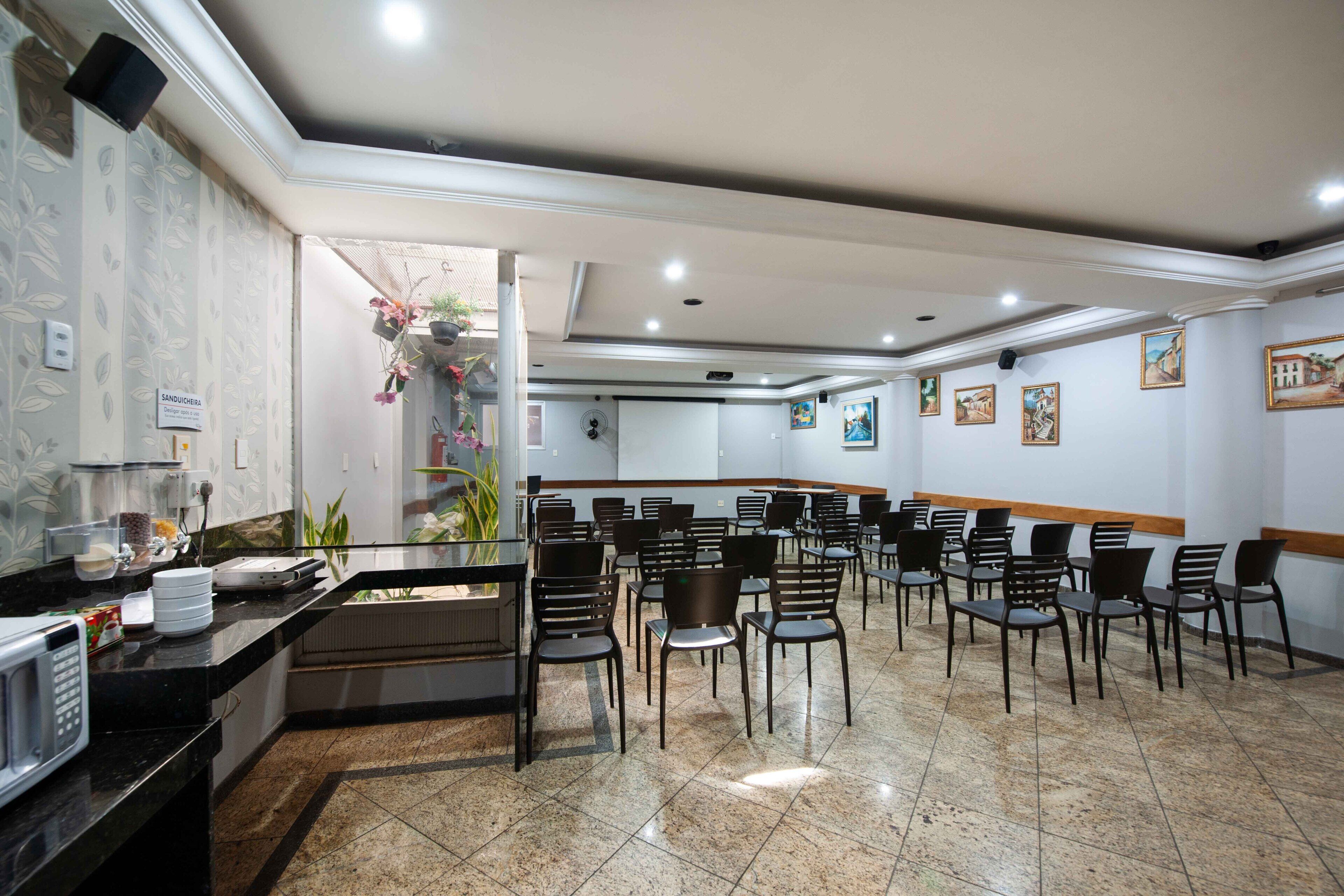 Un salón de conferencias con una larga mesa y sillas, una zona de cocina con electrodomésticos y una planta decorativa.