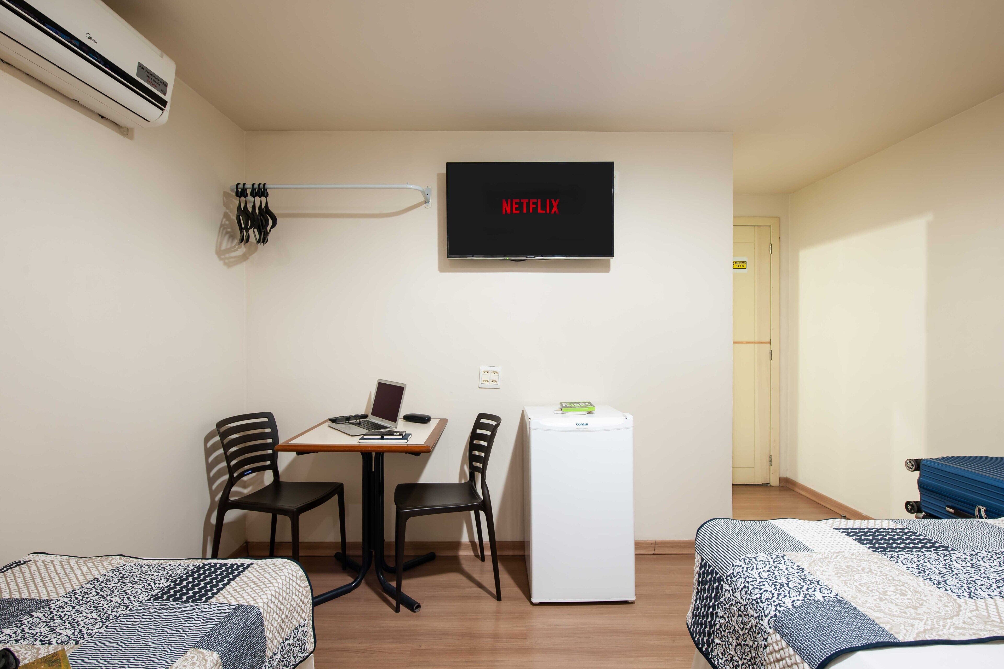 A room with a desk, two chairs, a laptop, a TV displaying 'NETFLIX', a mini-fridge, and a bed with patterned bedding.