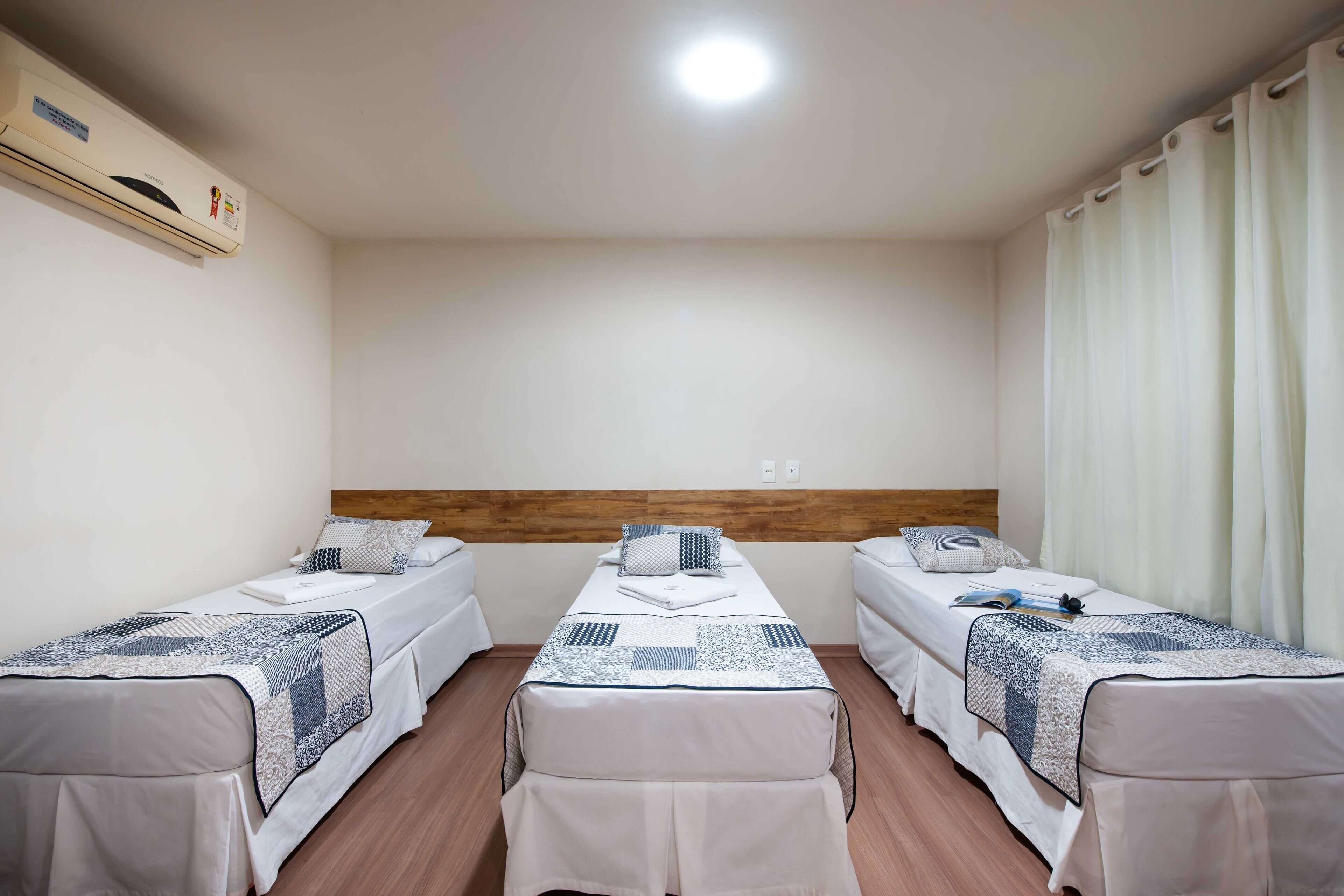 Quarto de hotel com duas camas de solteiro, cabeceira de madeira, ar condicionado e janela com cortinas.