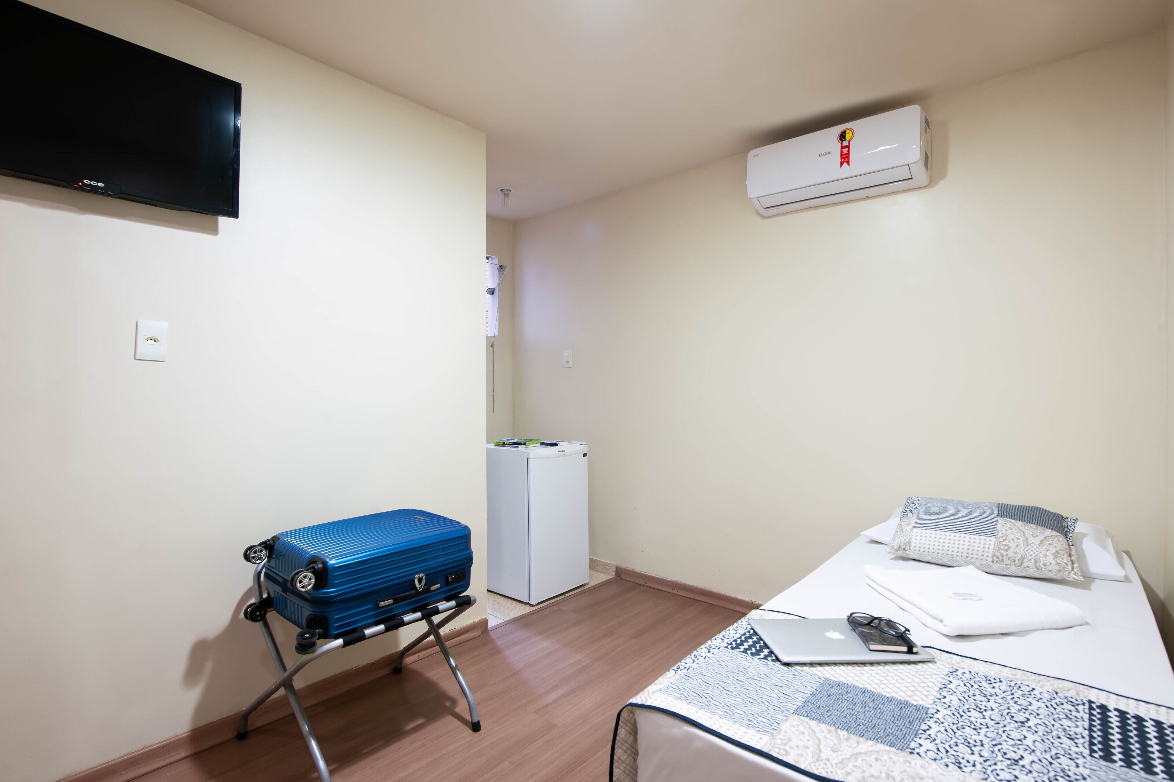 Um quarto pequeno com uma cama, uma escrivaninha, uma mala azul, um ar condicionado branco e uma TV na parede.