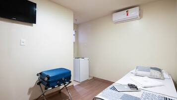 Um quarto pequeno com uma cama, uma escrivaninha, uma mala azul, um ar condicionado branco e uma TV na parede.