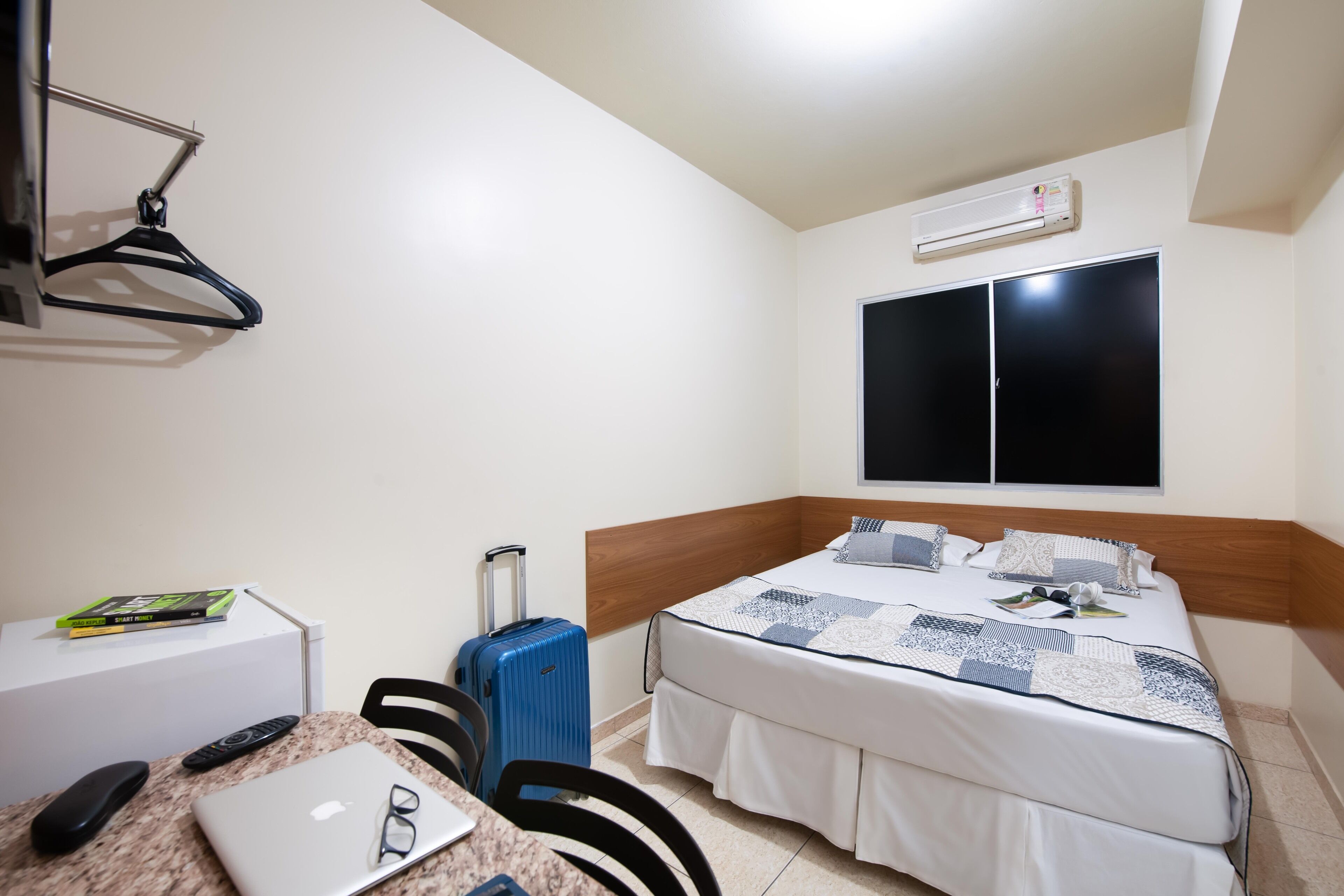 Quarto de hotel com cama, escrivaninha, laptop e controles remotos, mala e um cabideiro.