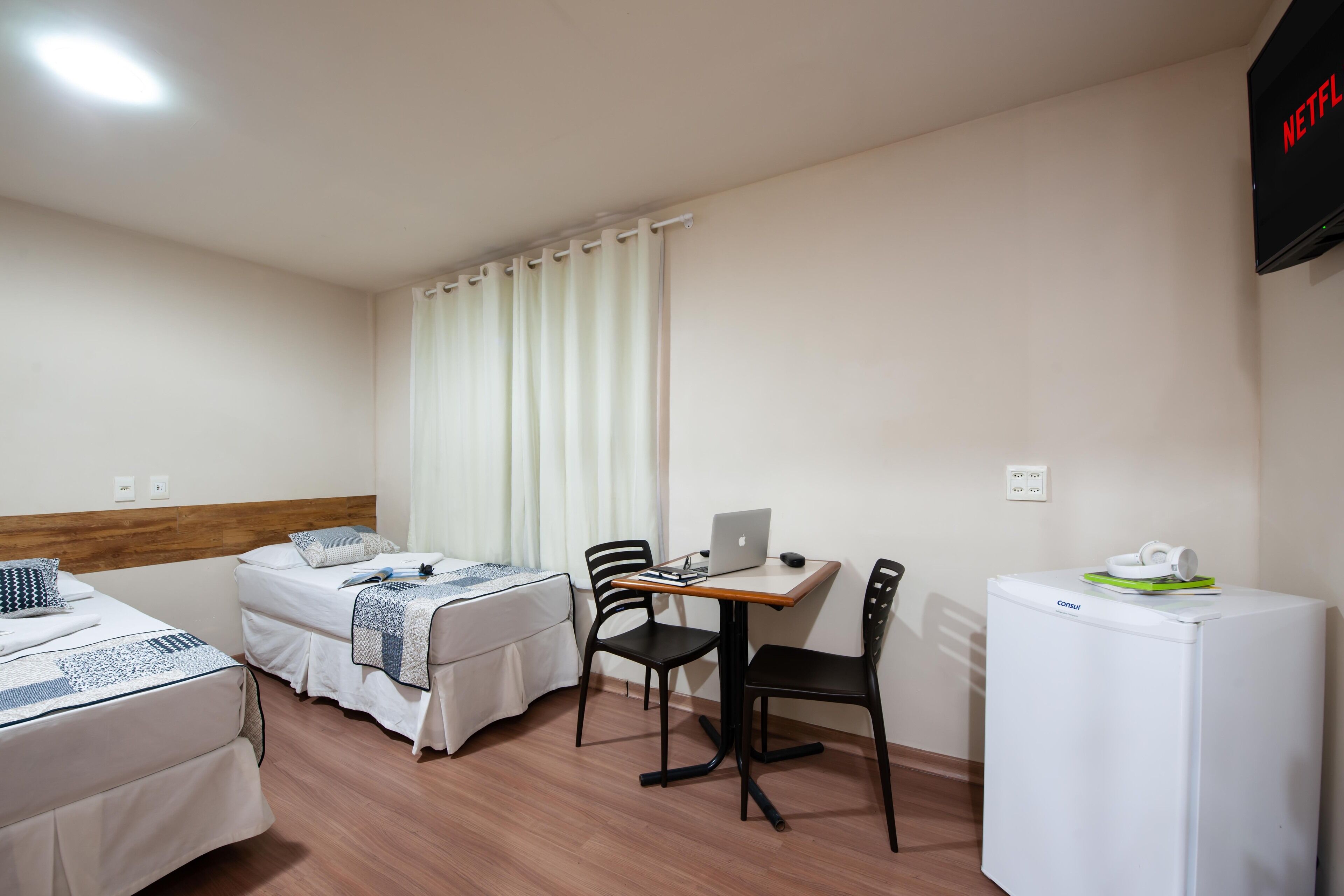 Habitació d'hotel amb dos llits, una escriptori amb un ordinador portàtil, cadires i un frigorífic.