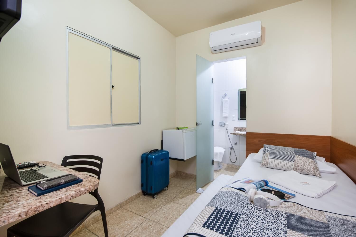 A hotel room with a bed, a desk with a laptop, a suitcase, a shower, and an air conditioning unit.