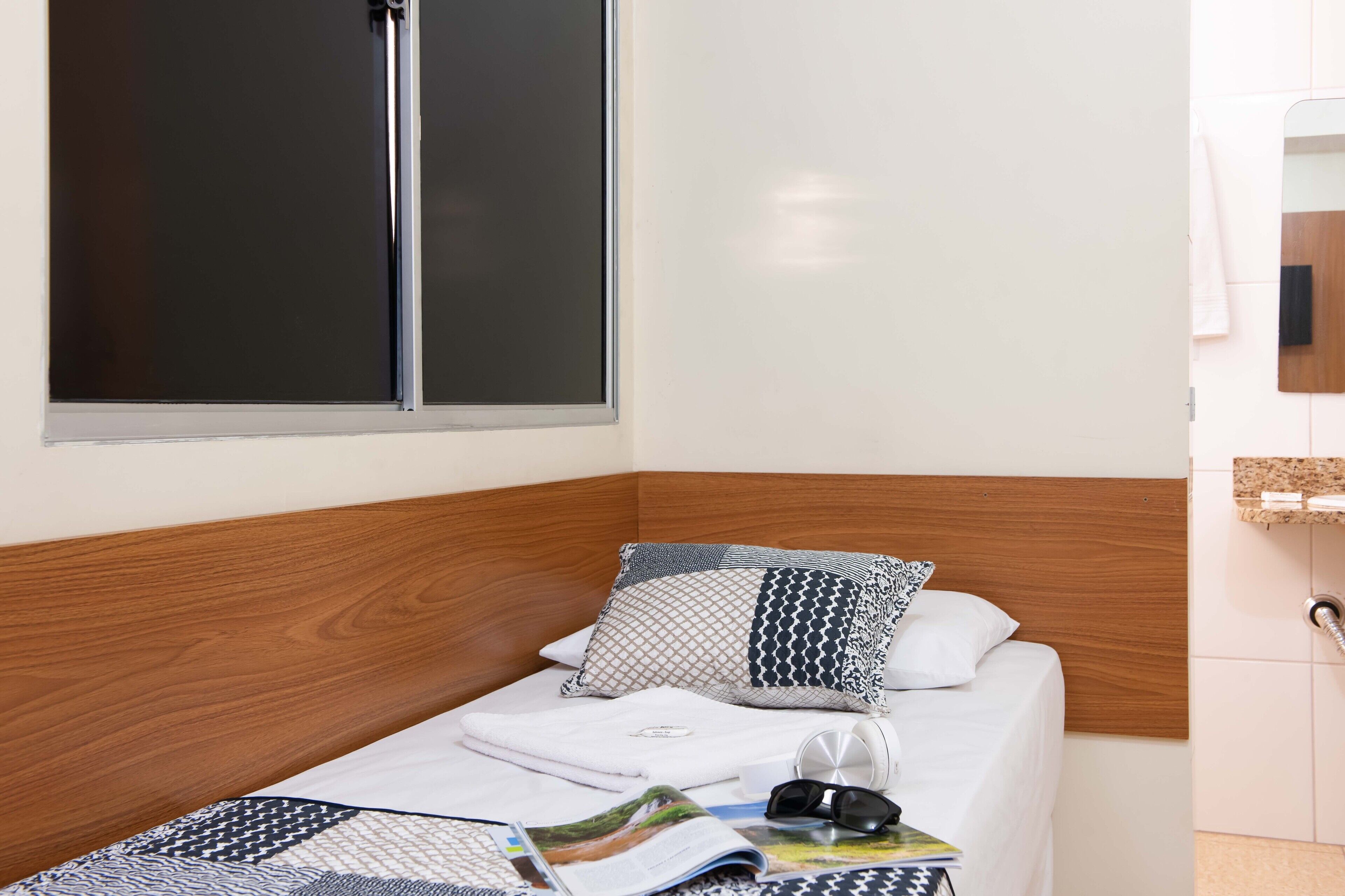 A small, neatly made bed with a wooden headboard, a window with blinds, and a small bedside table with a magazine, a pair of sunglasses, and a cup.