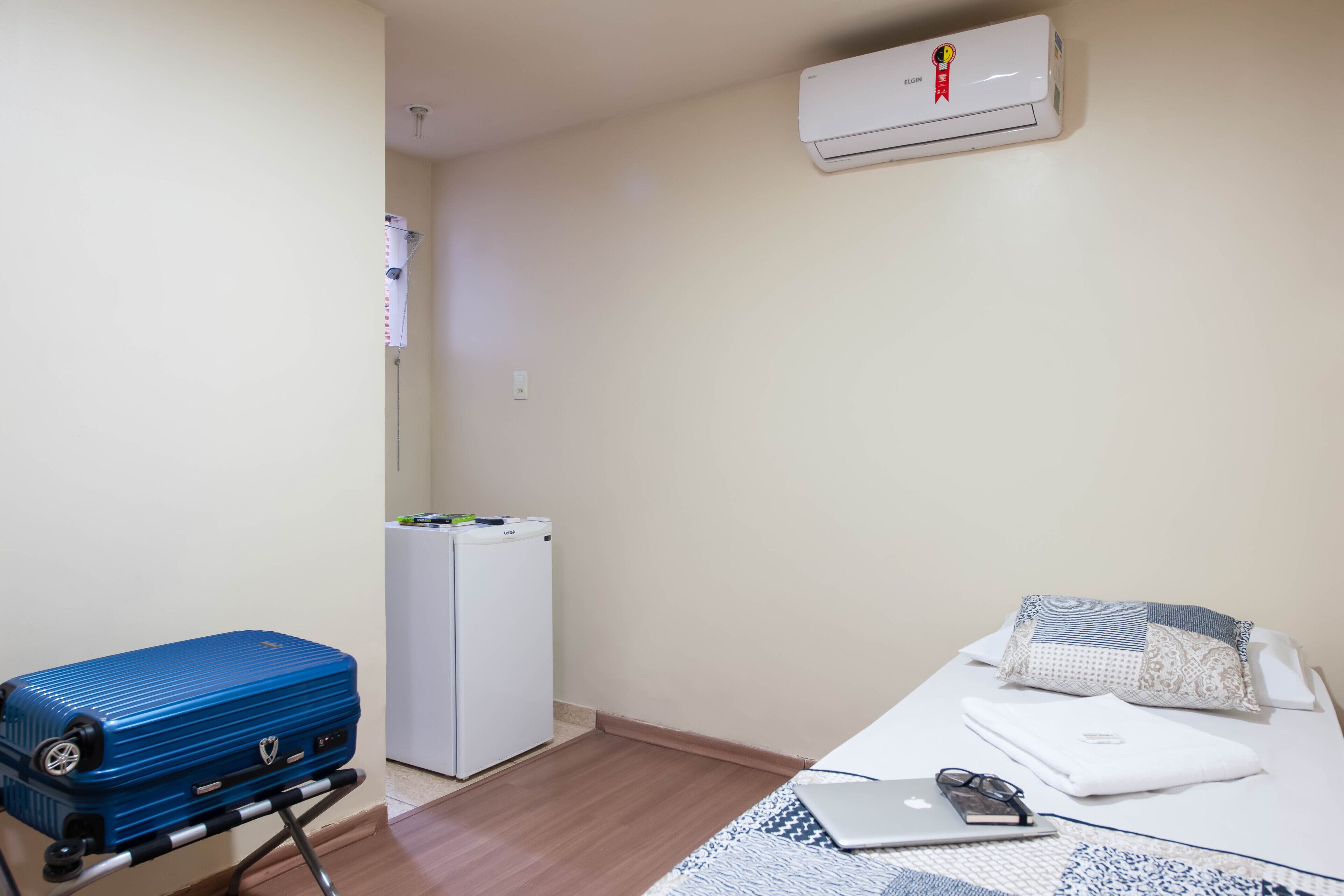 A room with a bed, a laptop, a pair of glasses, a blue suitcase, and a white refrigerator.