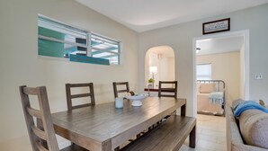 Un comedor con una mesa de madera, sillas y un plato de comida. Se ve una ventana con vistas a un edificio y un cartel sobre la ventana.