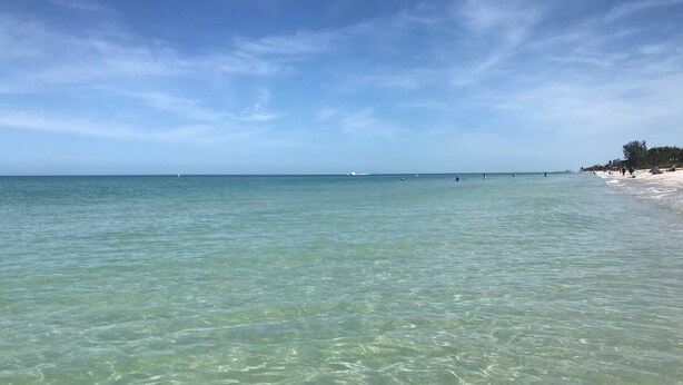Una playa con aguas claras y poco profundas, embarcaciones a lo lejos y algunas personas en la distancia.