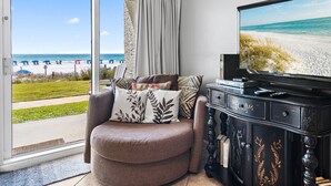 A living room with a large TV, a sofa with patterned cushions, a dark wooden cabinet, and a view of a beach through a sliding glass door. - Beach Service Included! | 1BR Windancer 111 | Beachfront | Heated Pool | 1st Flo (Miramar Beach)