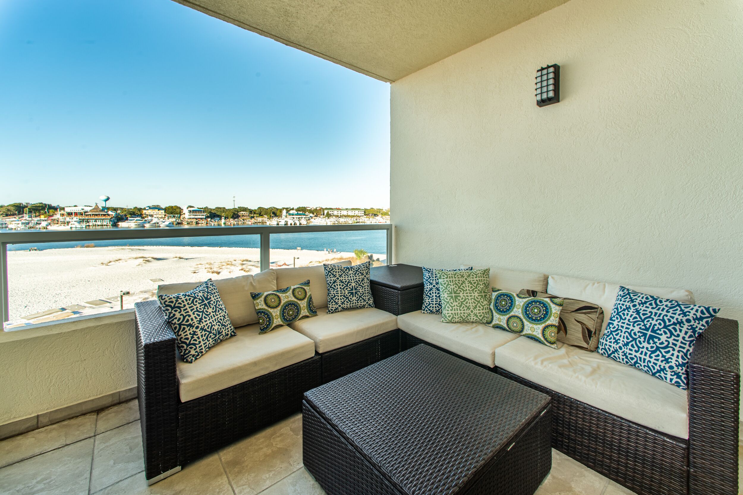A balcony with a wicker sofa and ottoman, a view of the beach, and a clear sky.