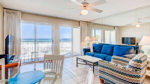 Ein geräumiges Wohnzimmer mit großem Fenster mit Blick auf einen Strand, einem blauen Sofa, einem weißen Sessel und einem Glastisch.