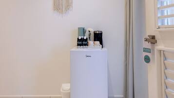 A white minibar with a macrame wall hanging, a coffee machine, and a small trash bin.
