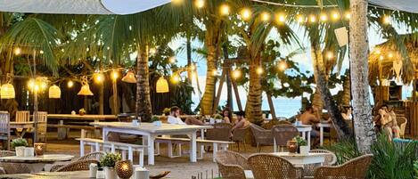 An outdoor dining area with tables and chairs, string lights, and palm trees.