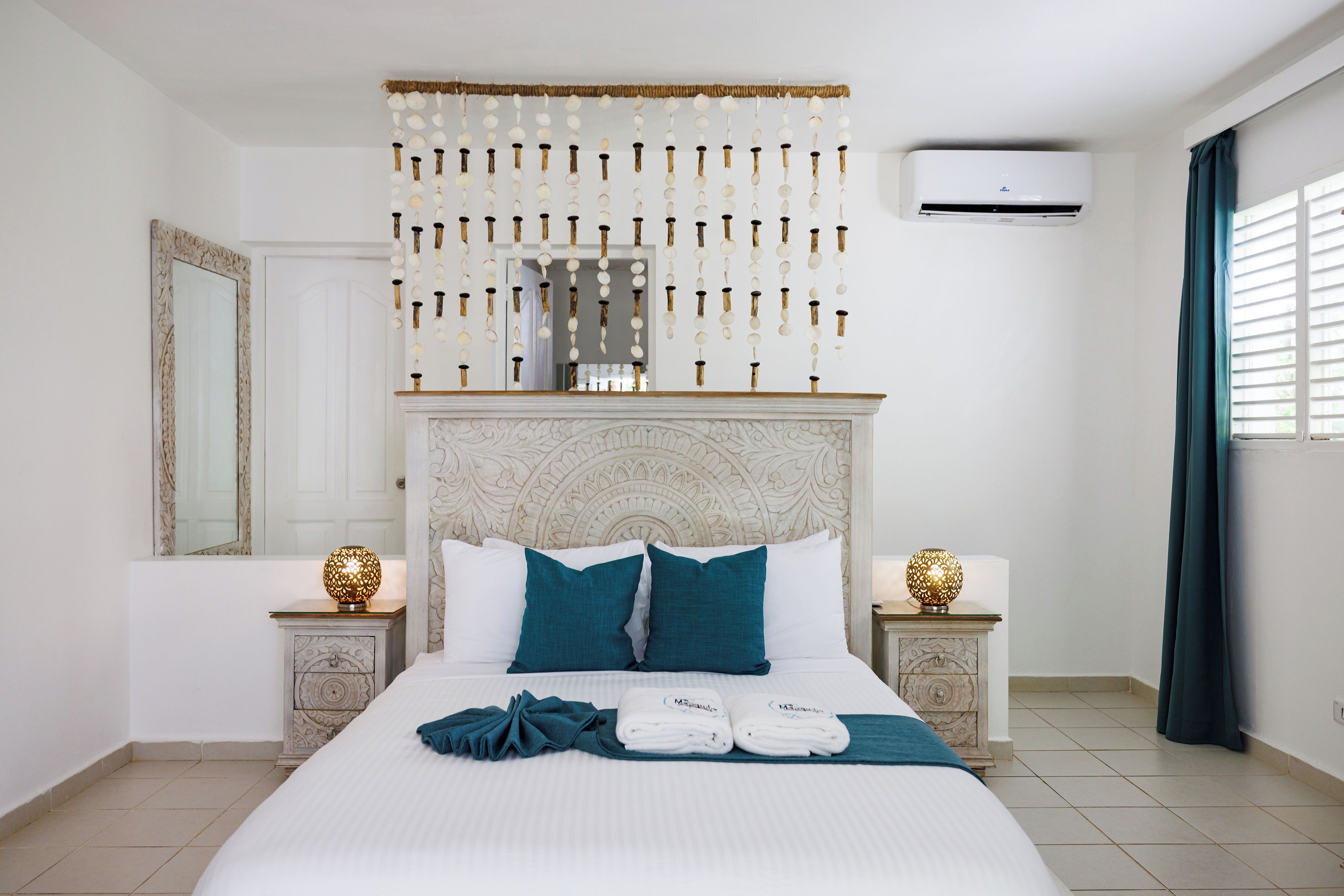 Een hotelkamer met een bed, twee nachtkastjes, een decoratief hoofdbord, een airconditioning en een raam met gordijnen.