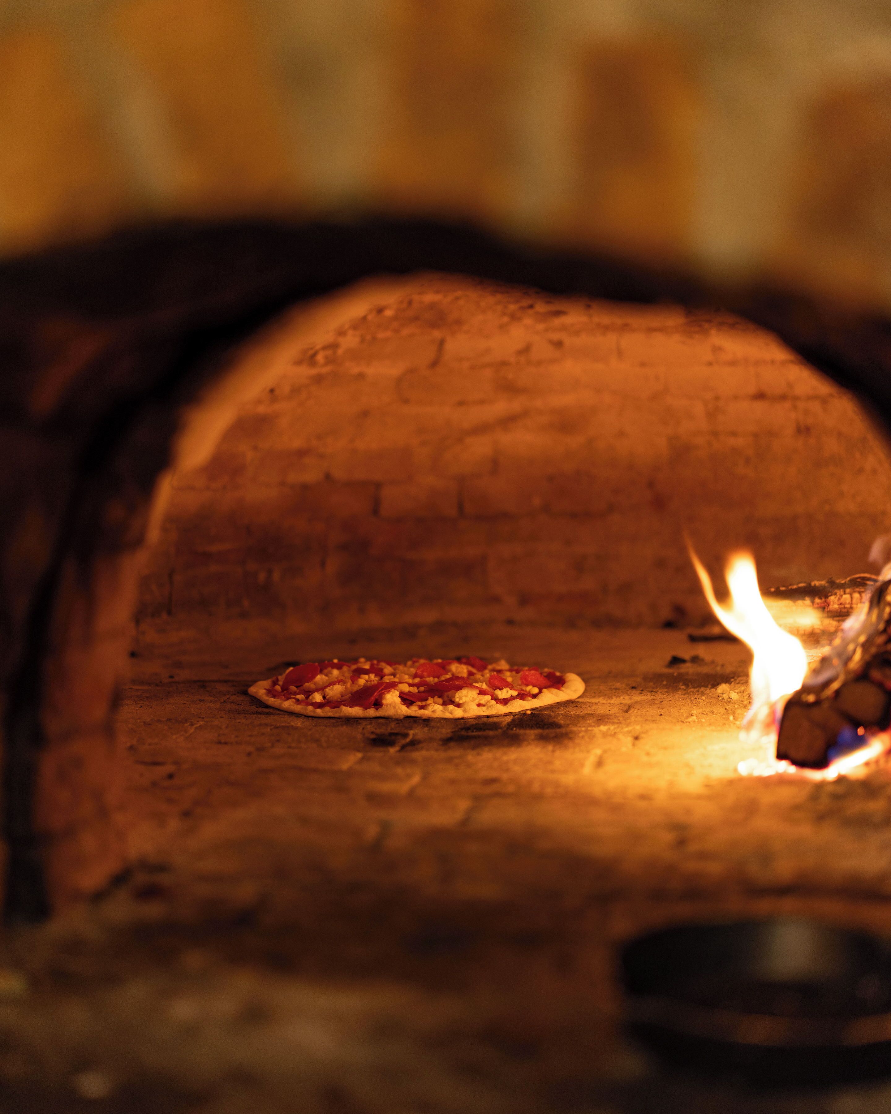A pizza oven with a pizza inside, a fire burning, and a brick wall in the background.