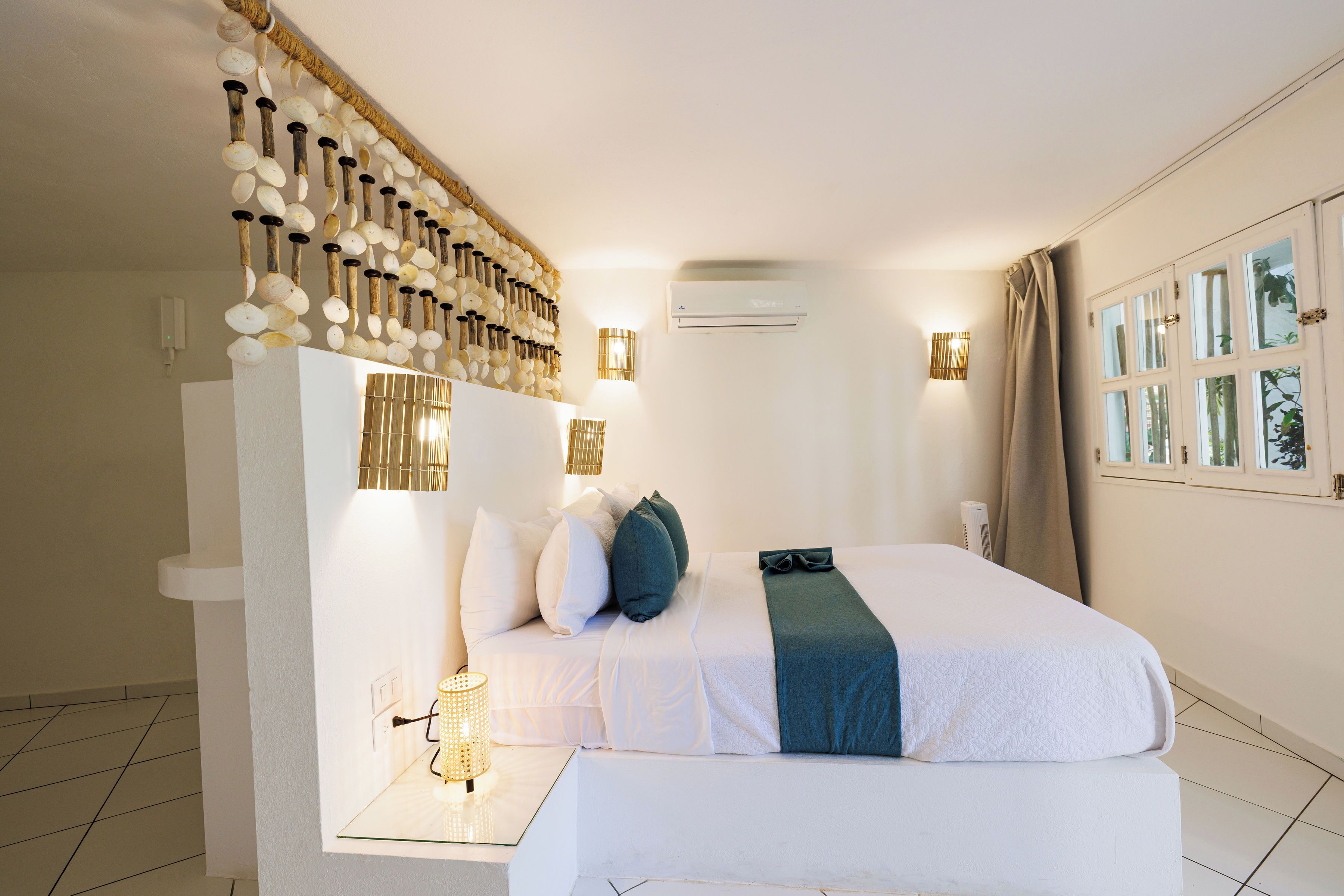A hotel room with a bed, a wall-mounted light fixture, an air conditioning unit, and a window with curtains.