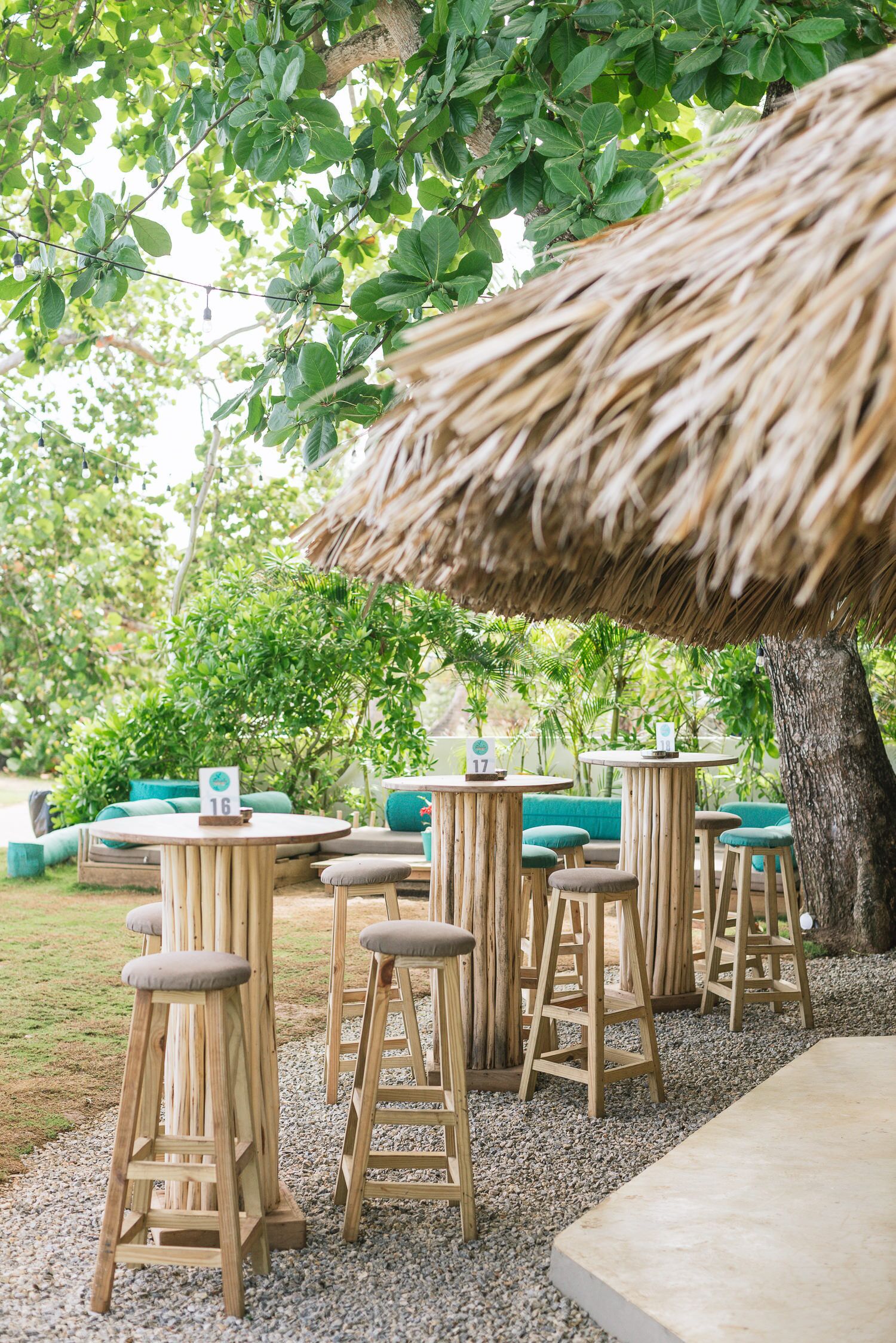 Un área de estar al aire libre con ambiente tropical, con taburetes y mesas de madera, rodeada de follaje y un paraguas de paja.