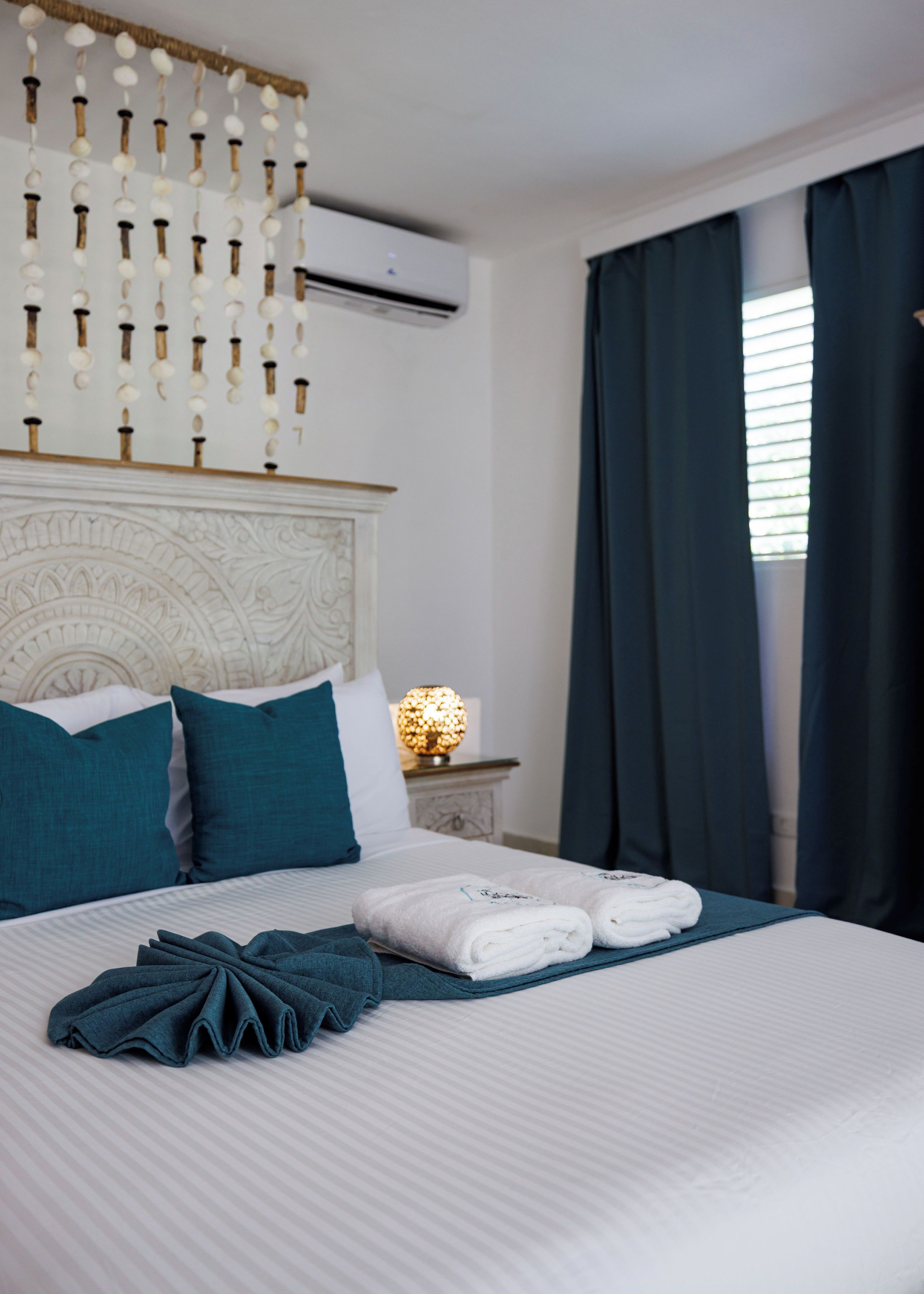 A neatly made bed with white linens, teal pillows, and a folded towel on a striped bedspread.