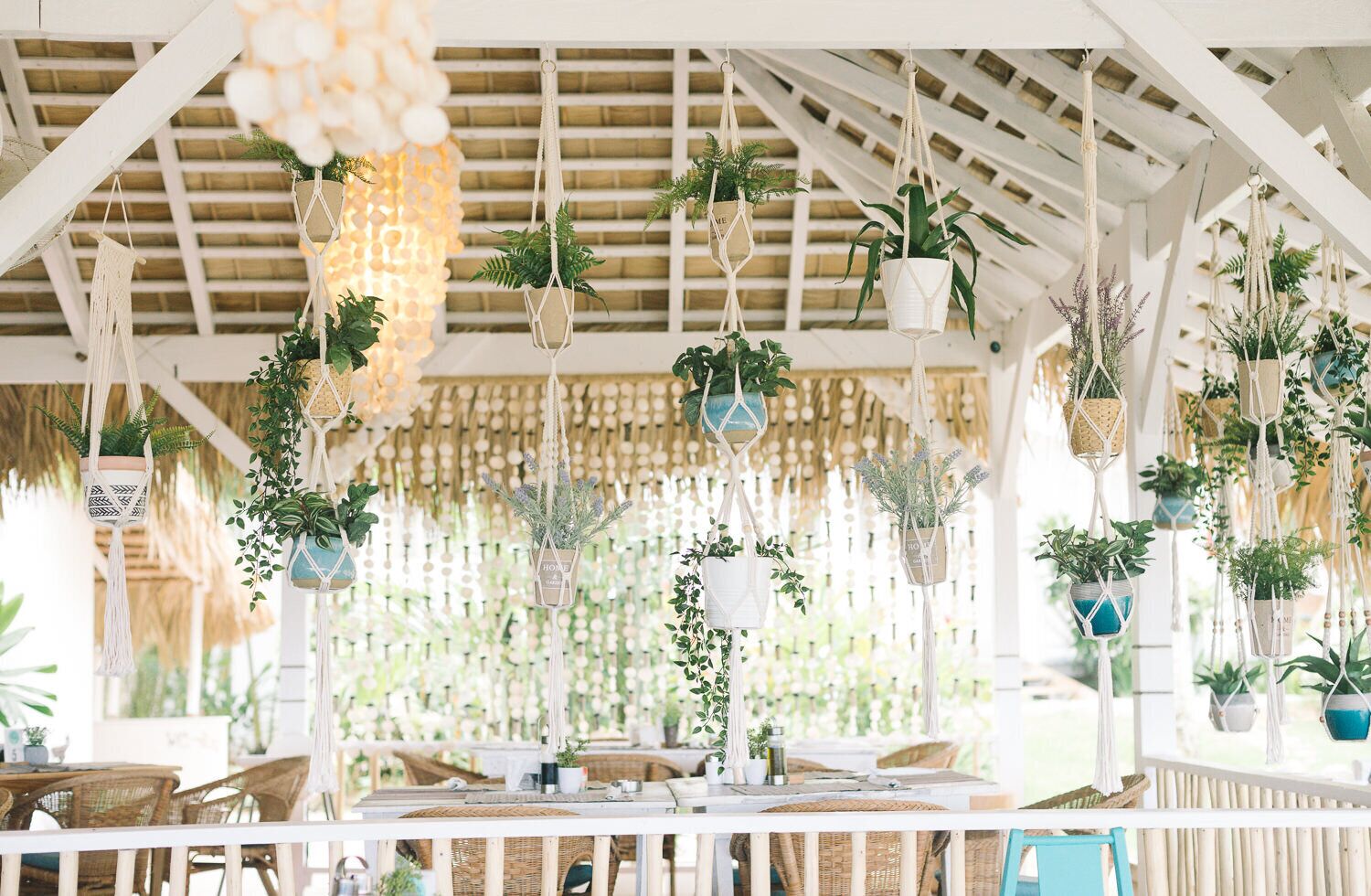 Uma área de estar ao ar livre coberta, com várias mesas e cadeiras, decorada com plantas pendentes e peças de macramé.