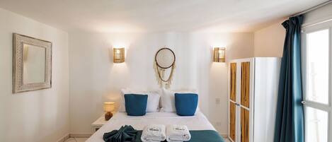 A hotel room with a bed, a bedside table, a wall-mounted mirror, and a view of the outside.
