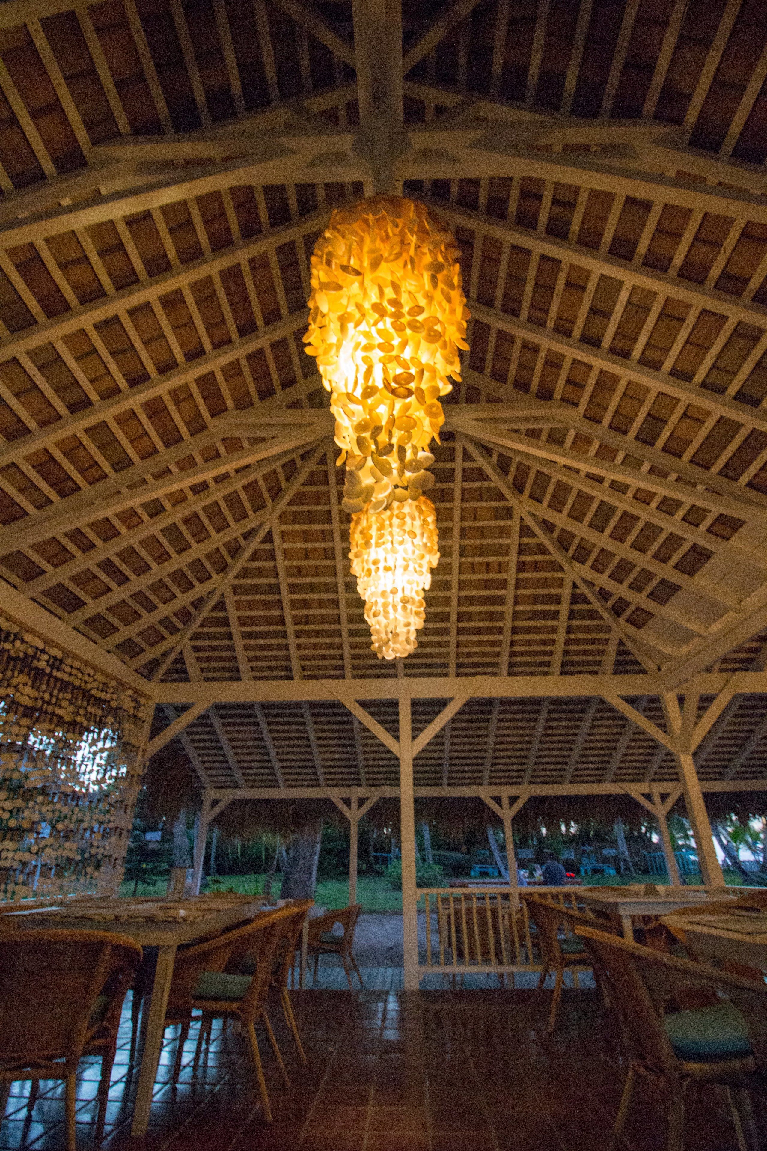 Een eetruimte binnenshuis met een hoge plafond en een hangende kroonluchter. Tafels en stoelen staan klaar voor een maaltijd.