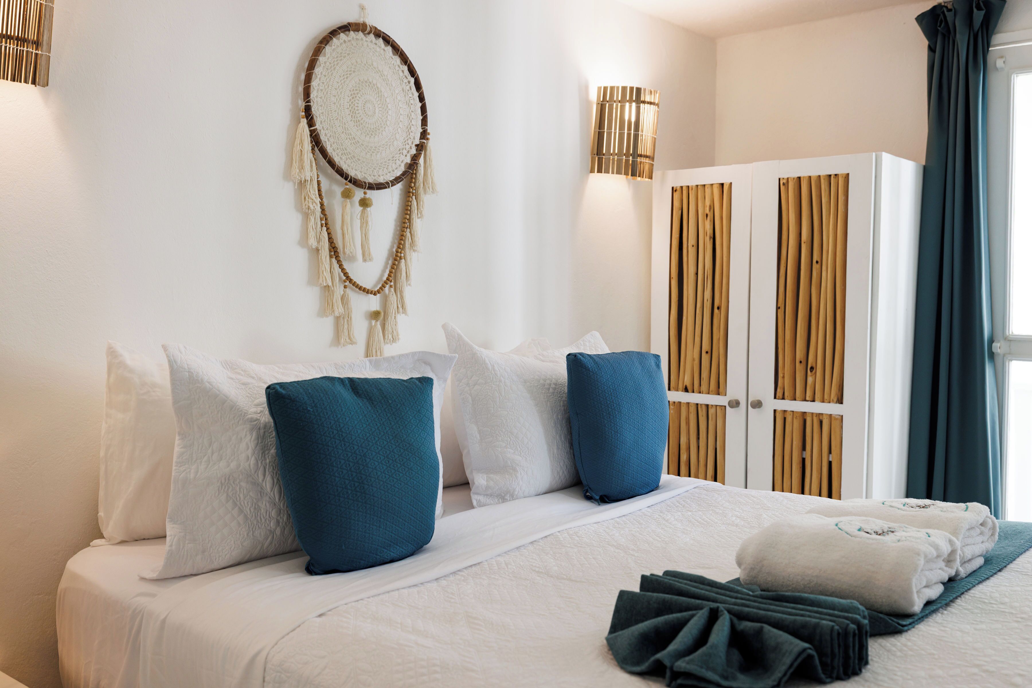 A neatly made bed with white and blue pillows, a dreamcatcher on the wall, and a wardrobe with wooden doors.