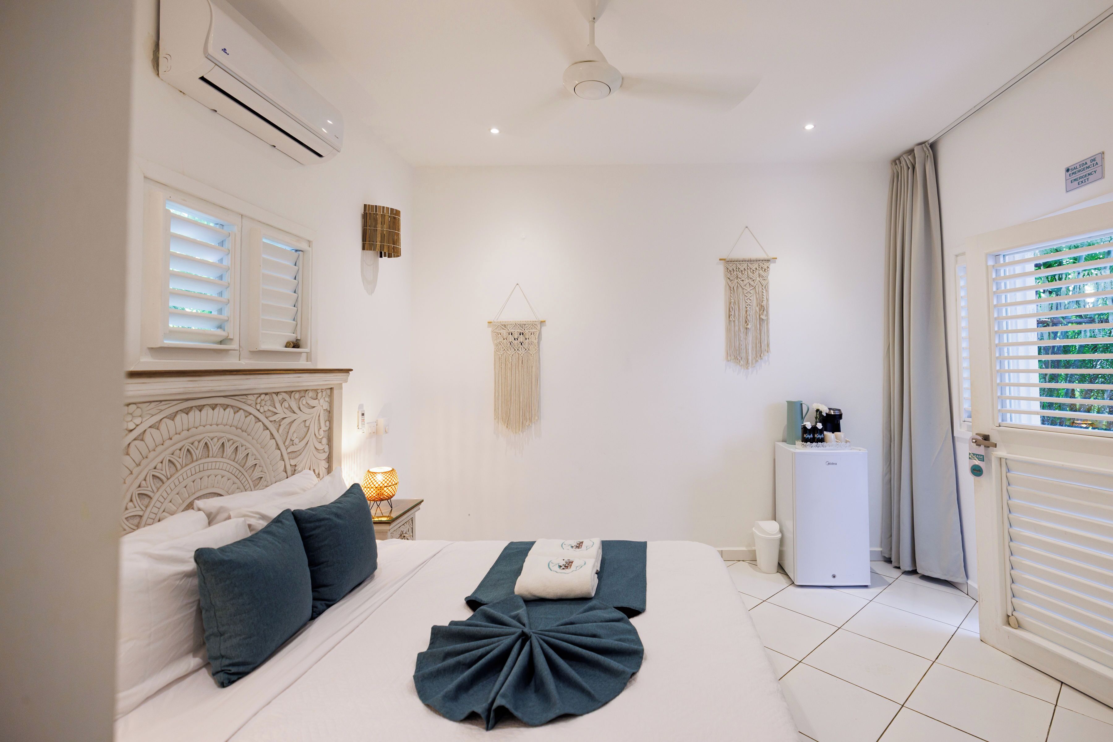 A bedroom with a bed, bedside table, air conditioning unit, decorative wall hangings, and a door with curtains.