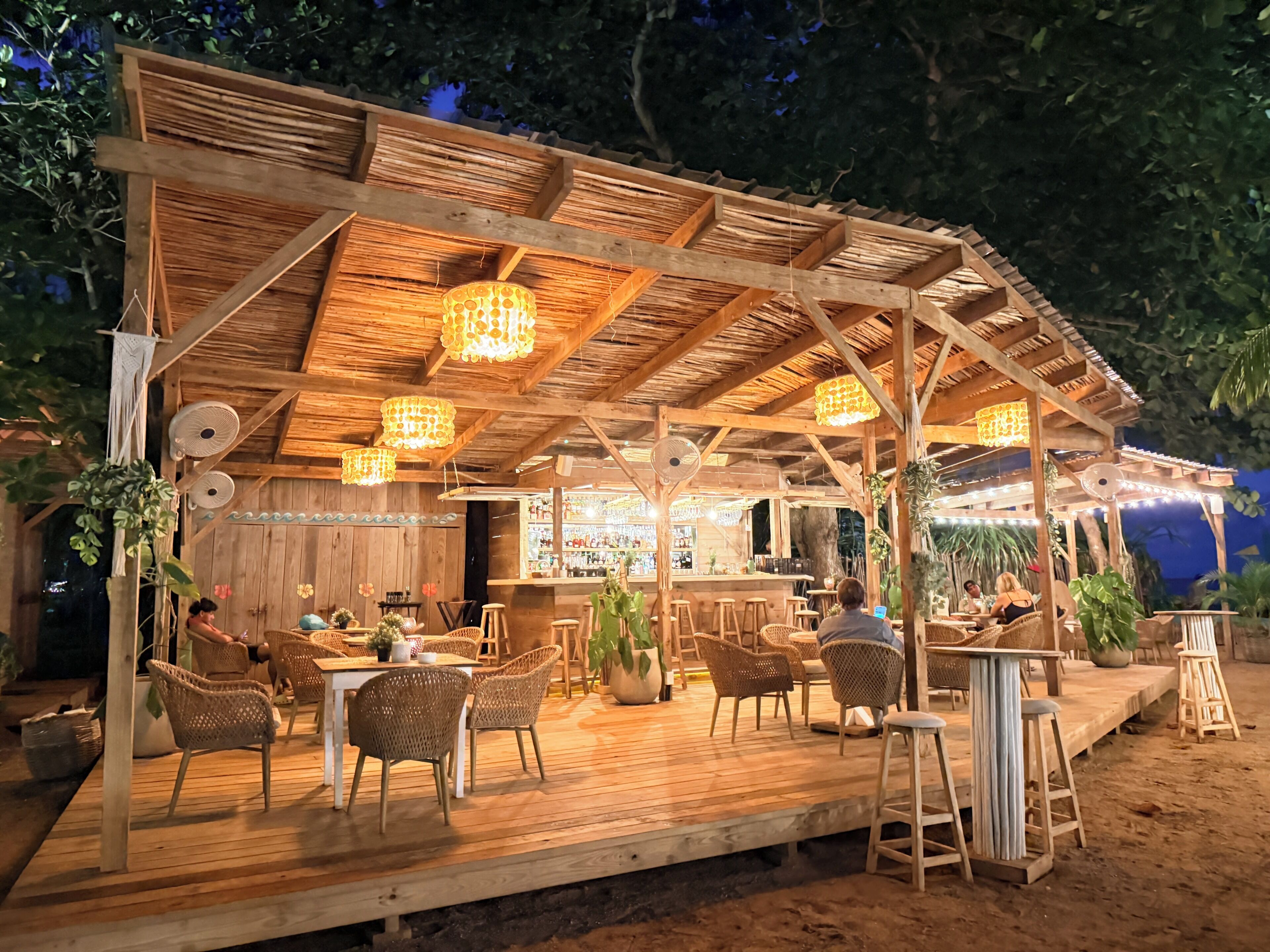 An outdoor wooden structure with a bar, wicker furniture, and hanging lights.