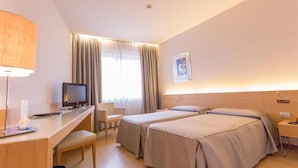 Quarto de hotel com duas camas, uma mesa com computador, uma cadeira e uma luminária.