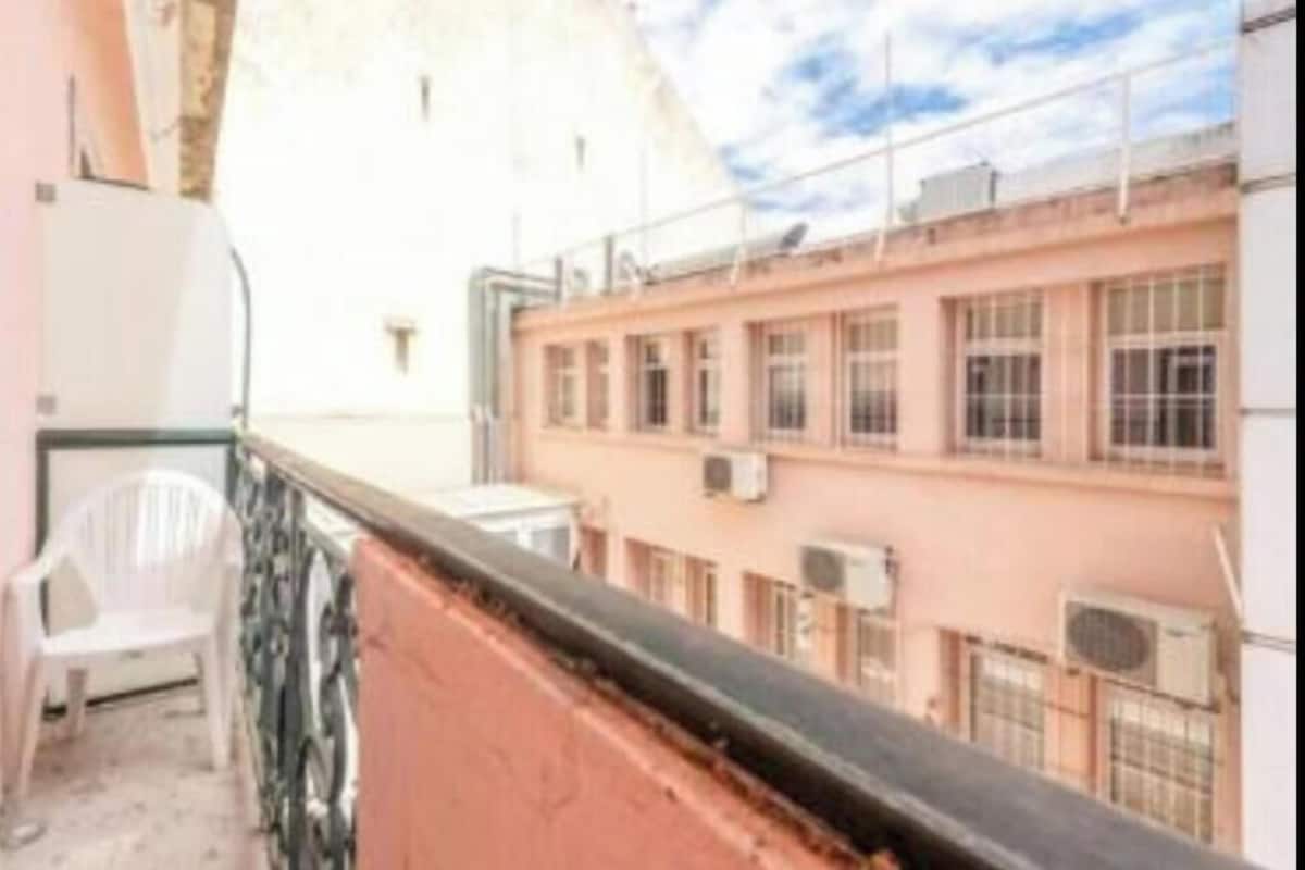 A balcony with a white plastic chair, a pink building with multiple windows, and an air conditioning unit.