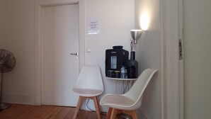 A room with a white door, a standing fan, a white chair, a small table with a coffee maker and water bottles, and a wall-mounted lamp. - Happy @ Chiado (Lisbon)