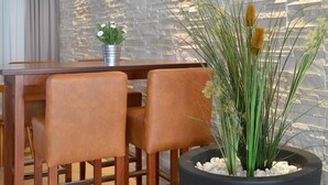 Un comedor moderno con una mesa de madera y dos sillas de cuero marrón, una planta en maceta con flores blancas, y plantas verdes altas con flores amarillas.