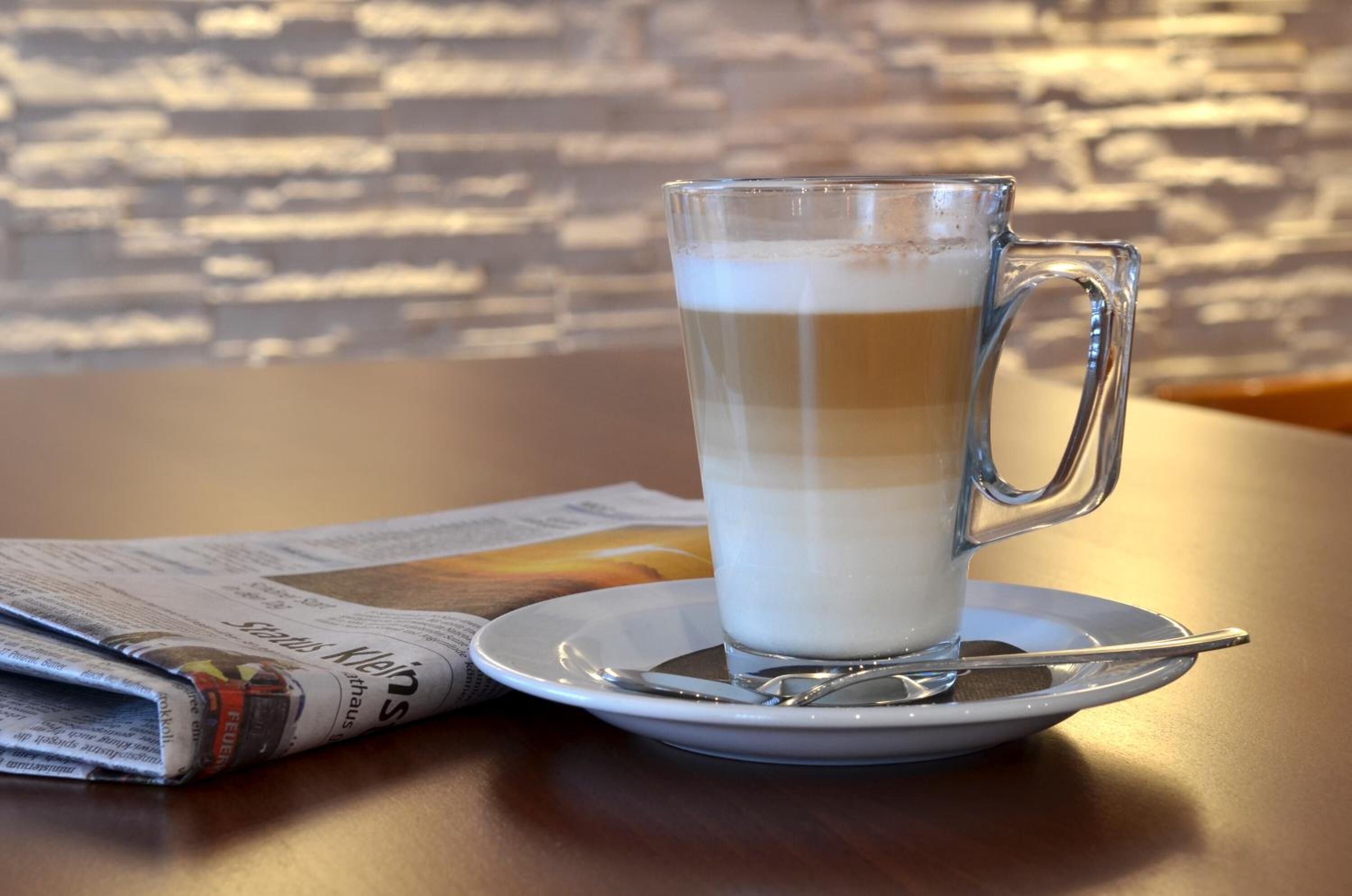 Una taza de vidrio transparente con asa sobre un platillo, colocada sobre una mesa con un periódico a un lado.