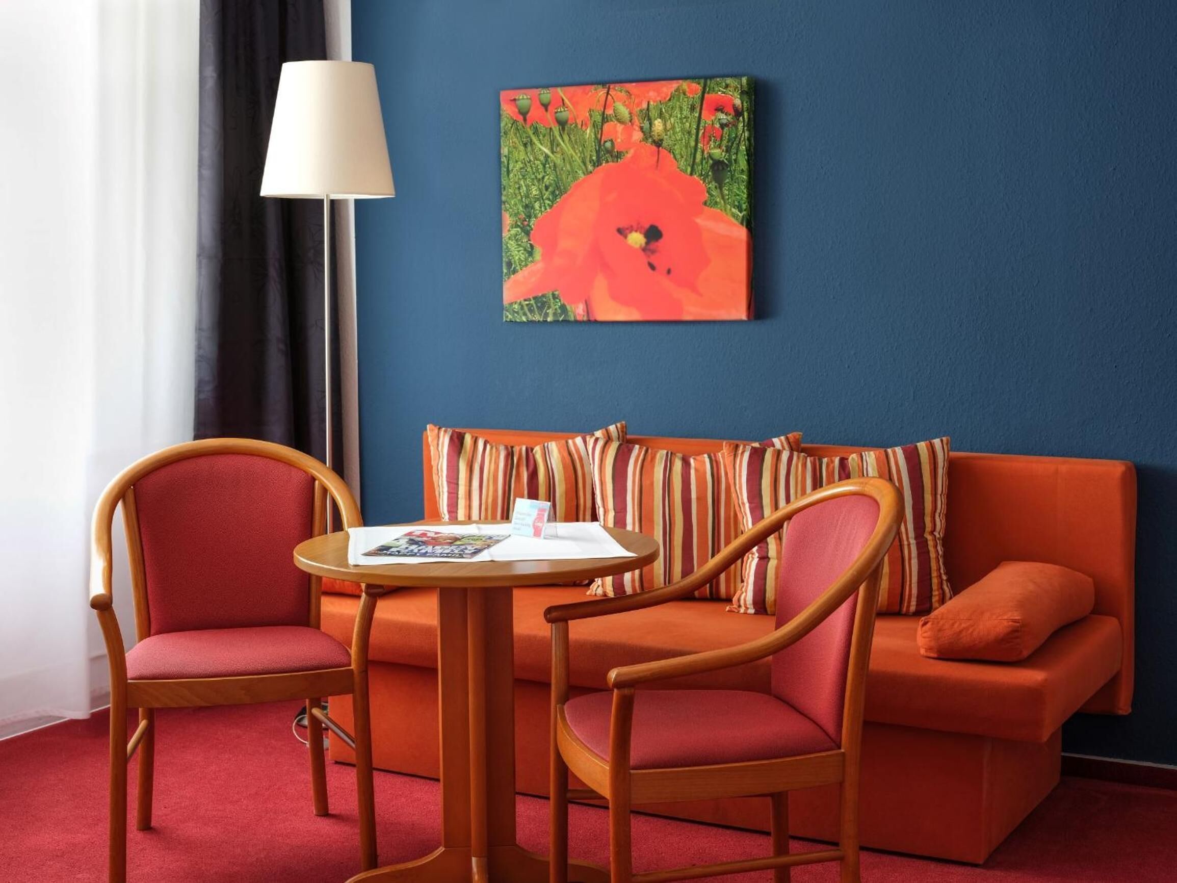 A room with a blue wall, a red sofa, two wooden chairs, a small round table, and a lamp.